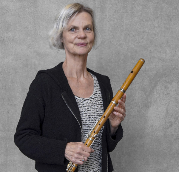 Person steht vor einer grauen Wand und hält eine Traversflöte aus hellem Holz; trägt ein strukturiertes Oberteil und eine dunkle Jacke.