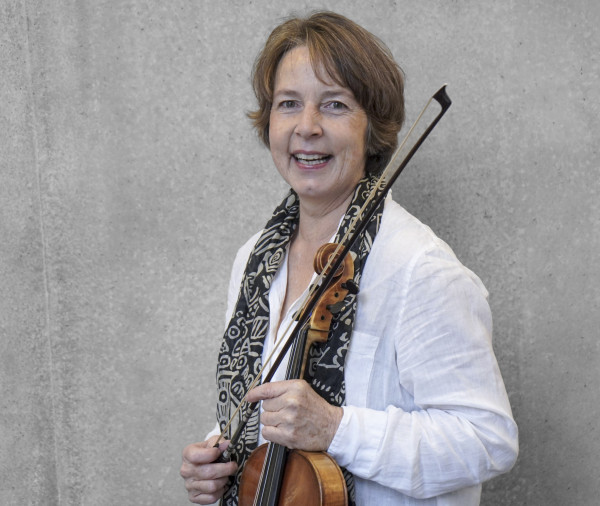 Person steht vor einer grauen Wand und hält eine Bratsche mit Bogen; trägt ein helles Hemd und einen gemusterten Schal.