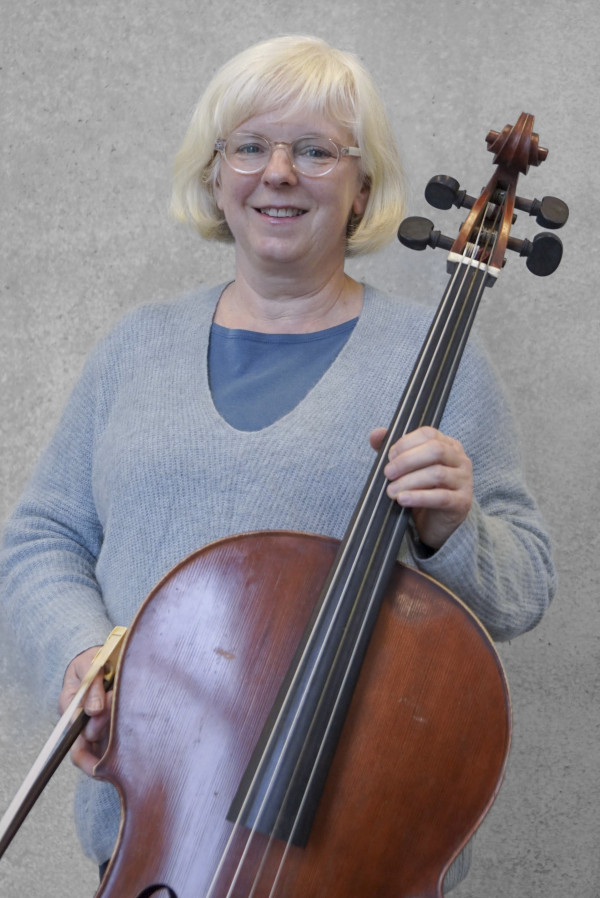 Person hält ein großes Streichinstrument, ein Cello, vor einem neutralen grauen Hintergrund; die Person trägt einen hellen Pullover und hält Bogen und Instrument in Spielposition