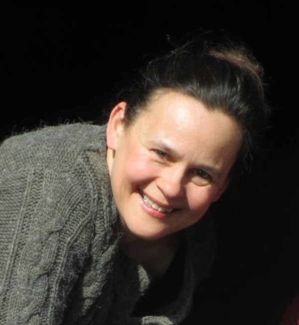 Person befindet sich vor dunklem Hintergrund und trägt einen dicken, grob gestrickten Pullover; der Oberkörper ist leicht nach vorn geneigt und die Beleuchtung fällt punktuell auf die Kleidung.