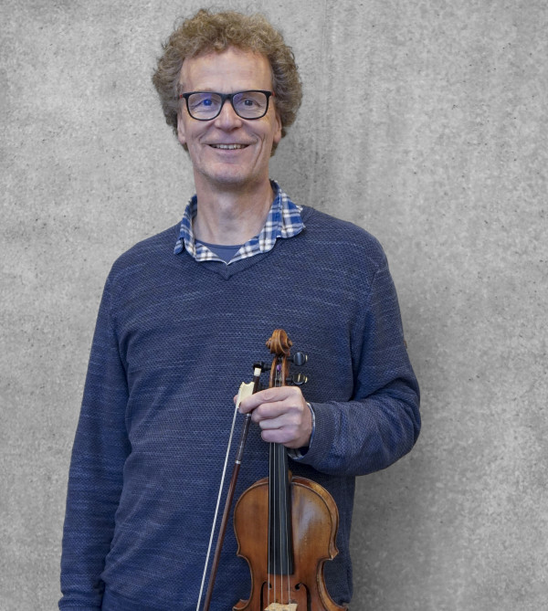 Person steht vor einer grauen Wand und hält eine Geige mit Bogen locker in der Hand; trägt einen blau melierten Pullover über einem karierten Hemd.