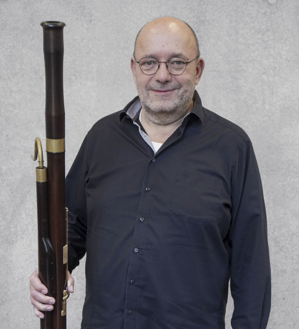 Person steht vor einer hellgrauen Wand und hält ein großes historisches Blasinstrument, ein Fagott mit Holz- und Messingelementen; dunkel gekleidet und frontal zur Kamera positioniert.