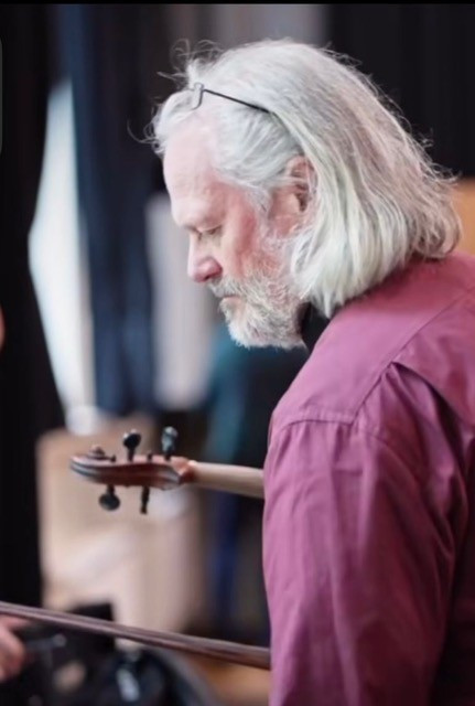 Person steht in einem Innenraum mit weichem Bühnen‑ oder Probenlicht und hält eine Geige seitlich am Hals des Instruments; trägt ein weinrotes Hemd und hat helles, schulterlanges Haar.