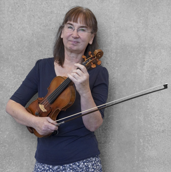 Person steht vor einer grauen Wand und hält eine Geige in Spielposition; trägt ein dunkles Oberteil mit halblangen Ärmeln und einen Rock mit Muster.