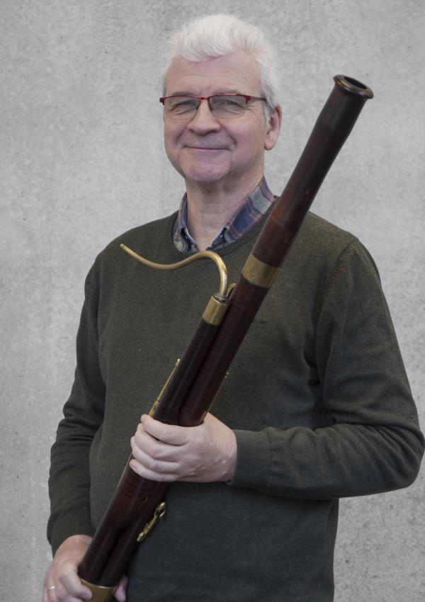 Person steht vor einer grauen Wand und hält ein großes historisches Blasinstrument aus Holz mit Metallrohr und Mundstück; trägt einen dunklen Pullover über einem karierten Hemd.