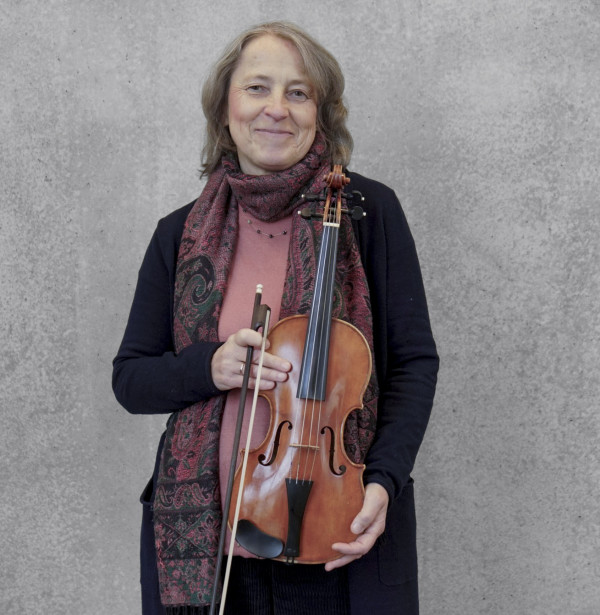 Person steht vor einer grauen Wand und hält ein Streichinstrument mit Bogen vor dem Körper; trägt einen dunklen Mantel sowie einen gemusterten Schal über einem rosafarbenen Oberteil.