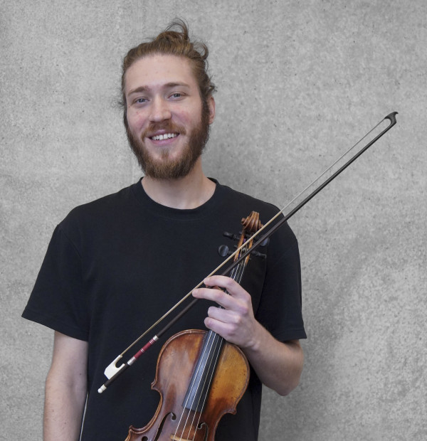 Person steht vor einer grauen Wand und hält eine Geige mit Bogen in der Hand; trägt ein dunkles T‑Shirt und hat das Haar zu einem Knoten gebunden.