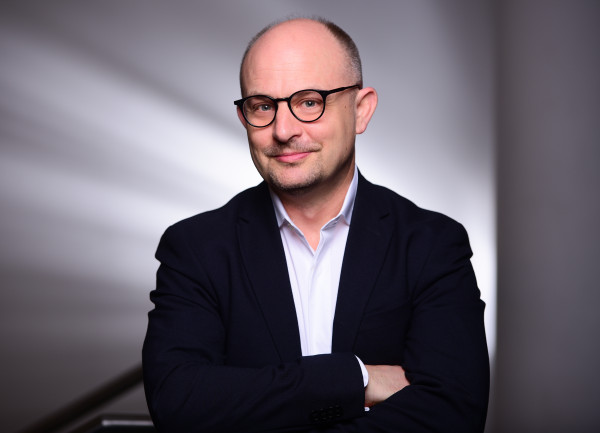 Person steht vor einem abstrakten, hellen Hintergrund mit diagonalen Lichtbahnen; trägt ein Jackett und ein helles Hemd und hat die Arme vor dem Körper verschränkt.