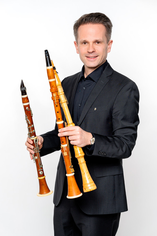 Person steht vor einem weißen Hintergrund und hält drei historische Holzblasinstrumente, Klarinetten, in beiden Händen; trägt einen dunklen Anzug.