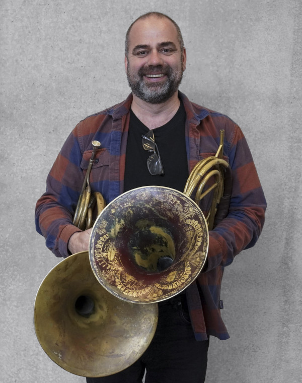 Person steht vor einer grauen Wand und hält zwei historische Blechblasinstrumente, Hörner, mit großen verzierten Schallbechern; trägt ein kariertes Hemd mit dunklem T‑Shirt darunter.