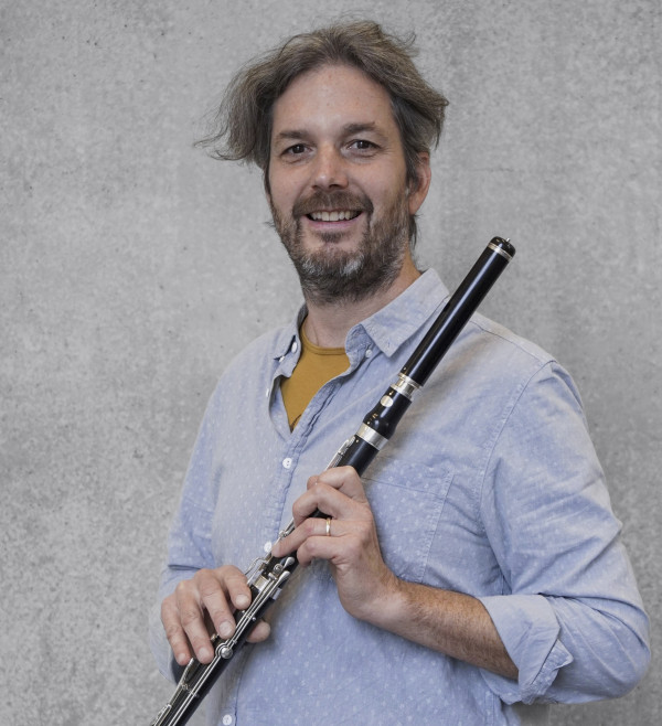 Person steht vor einer grauen, strukturierten Wand und hält eine schwarze Traversflöte mit silberfarbenen Klappen; trägt ein hellblaues Hemd und hält das Instrument mit beiden Händen in einer ruhigen Position.