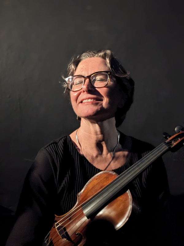 Person sitzt vor einem dunklen Hintergrund und hält eine Geige seitlich vor dem Oberkörper; Kleidung in dunklen Tönen und gerichtetes Licht, das Instrument und Teile der Kleidung betont.