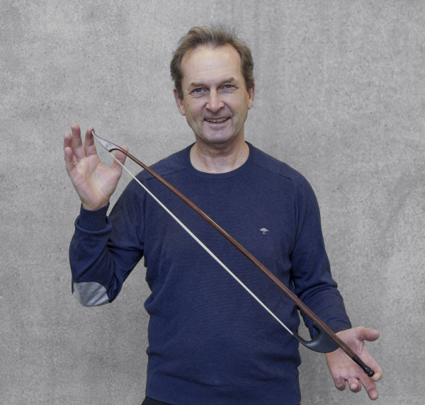 Person steht vor einer grauen Wand und hält einen langen Streichbogen locker in der Hand; trägt einen dunkelblauen Pullover.