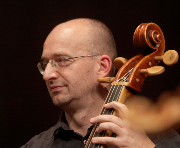 Person spielt ein großes Streichinstrument auf einer Bühne; der Fokus liegt auf dem Instrument und der Haltung der linken Hand am Griffbrett.