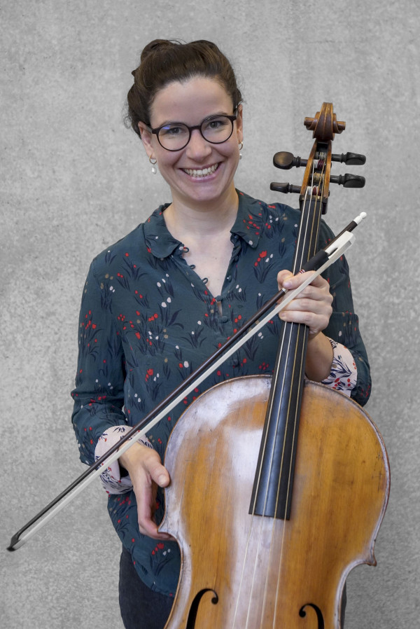 Person steht vor einer grauen Wand und hält ein Cello mit Bogen; trägt eine blumengemusterte, langärmlige Bluse.