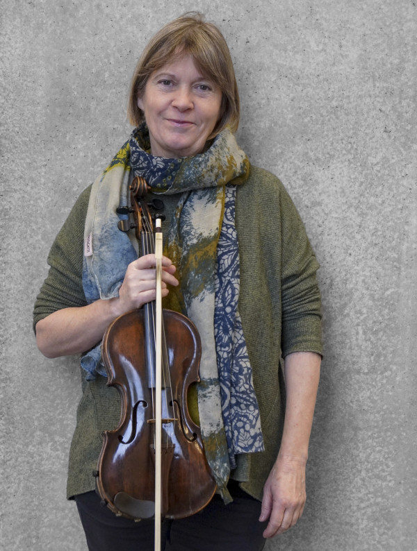 Person steht vor einer grauen Wand und hält eine Geige mit Bogen vor dem Körper; trägt einen grob gestrickten, grünen Pullover und einen großen mehrfarbigen Schal.