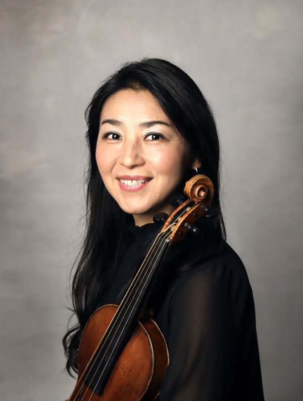 Person steht vor einem neutralen Hintergrund und hält eine Geige seitlich vor dem Oberkörper; trägt ein dunkles, leicht transparentes Oberteil und hat langes, dunkles Haar.