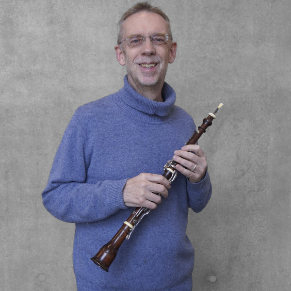 Person steht vor einer grauen Wand und hält ein schlankes Holzblasinstrument in beiden Händen; trägt einen strukturierten, hochgeschlossenen Strickpullover.