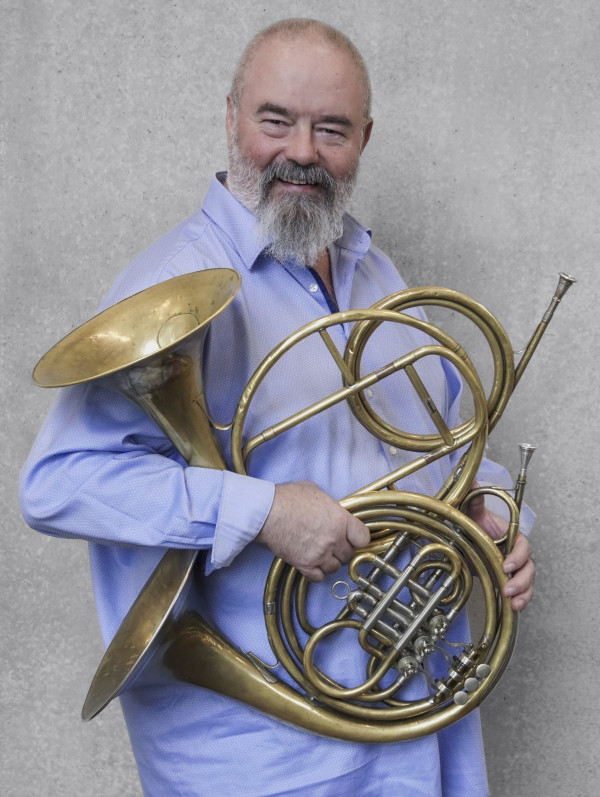 Person steht vor einer grauen Wand und hält zwei große historische Blechblasinstrumente mit verschlungenen Rohren und einem breiten Schallbecher, Hörner; trägt ein helles Hemd.