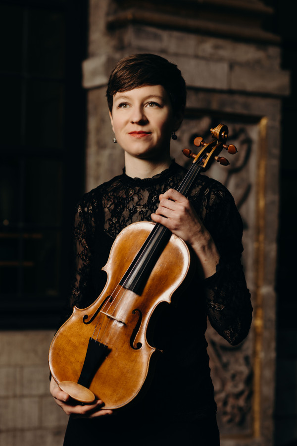 Person steht in einem historischen oder architektonisch verzierten Innenraum und hält eine Bratsche vor dem Oberkörper; trägt ein dunkles, fein strukturiertes Oberteil.