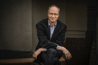 Fotografie eines sitzenden Mannes, der in die Kamera lächelt.