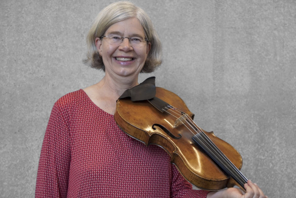 Person steht vor einer grauen Wand und hält ein Streichinstrument in Spielposition an der Schulter; trägt ein langärmliges Oberteil mit feinem Muster.