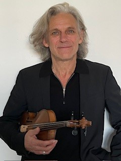 Person steht vor einer hellen Wand und hält eine Geige seitlich vor dem Körper; trägt eine dunkle Jacke mit Reißverschluss und hat das Instrument locker in beiden Händen.