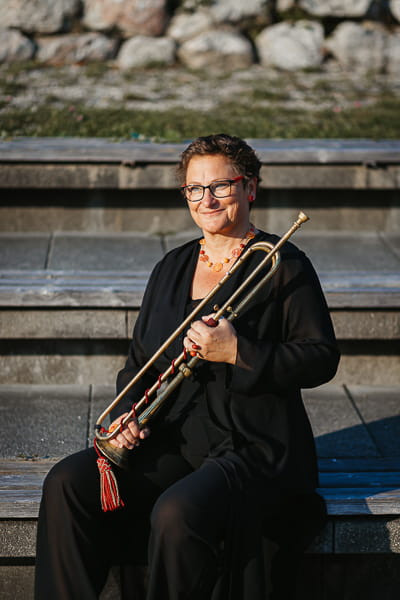 Person sitzt im Freien auf Steinstufen und hält eine historische Trompete mit roter Zierquaste; trägt ein dunkles Outfit und eine Halskette. Im Hintergrund sind Steine und Vegetation zu sehen.