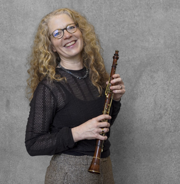 Alt‑Text 3 Person steht vor einer grauen Wand und hält ein hölzernes Blasinstrument, eine Oboe, mit mehreren Klappen; trägt ein dunkles, fein strukturiertes Oberteil und eine helle Hose.