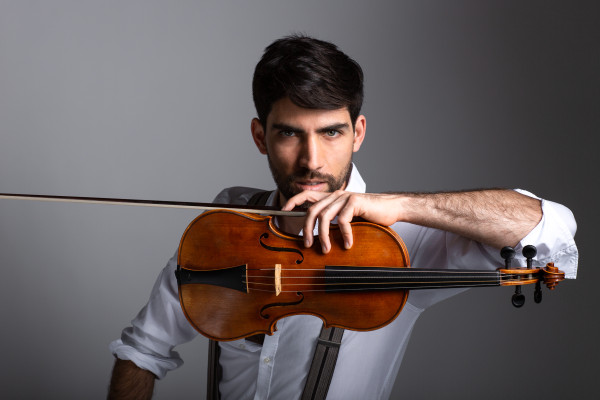 Person hält eine Geige in der Luft, der Arm liegt über dem Instrument; trägt ein helles Hemd mit hochgekrempelten Ärmeln und Hosenträgern vor einem neutralen Hintergrund.