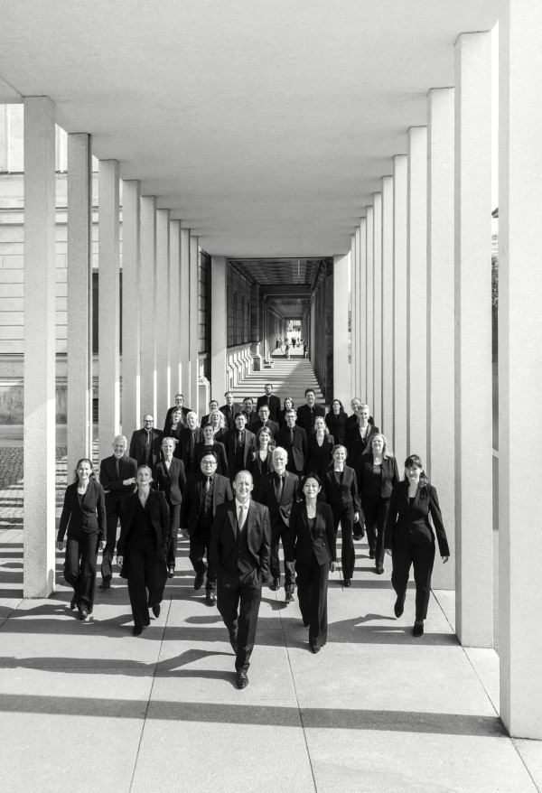 Eine Gruppe von Menschen in schicker Kleidung läuft auf den Fotografen des Bildes zu. Links und rechts sind Säulen zu sehen, die Sonne wirft Schatten. Eine schwarz-weiße Aufnahme