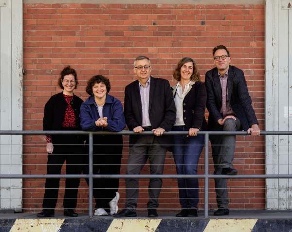 Fünf Personen stehen vor einer roten Backsteinwand hinter einem Geländer und lächeln.